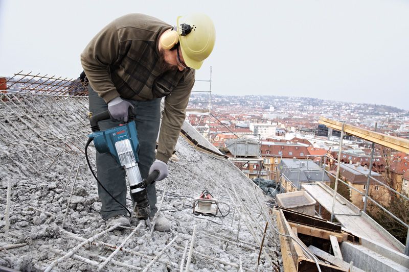 安全装備を身に着けた人が解体用ハンマーを使って屋上のコンクリートを破壊しています。.