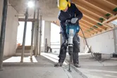 安全装備を身に着けた人が解体用ハンマーを使用してコンクリートの床を壊しています。.