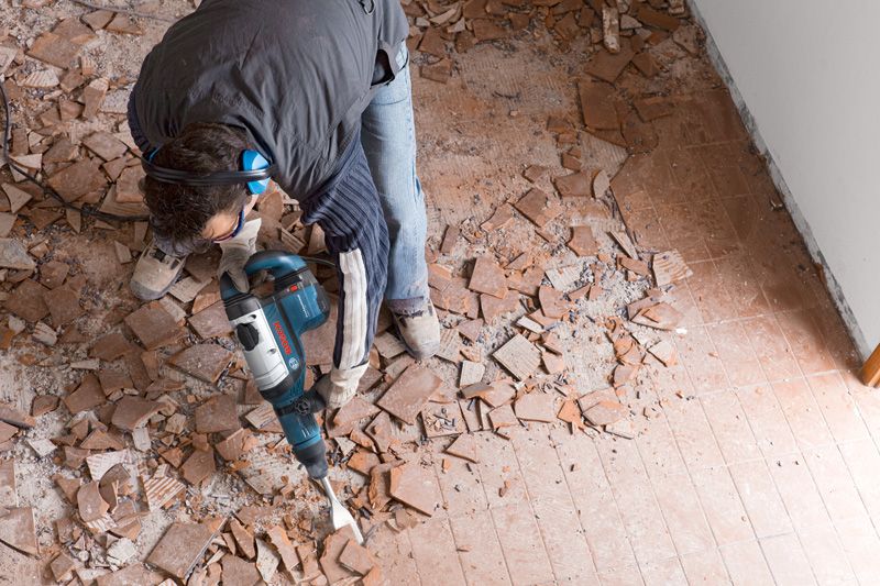 安全装備を身に着けた人が解体用ハンマーを使って床タイルを壊しています。.