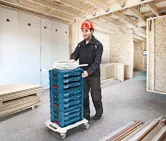 建設中の建物内で、安全装備を身に着けた人が、積み重ねられた工具ケースを台車に乗せて運んでいます。