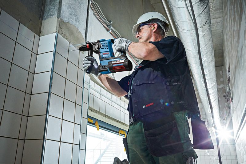 安全装置を装着した人が、コードレス回転ハンマーを使用してタイル張りの壁に穴を開けています。.