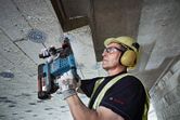 安全装備を身に着けた人がコードレス回転ハンマーでコンクリートの天井に穴を開けています。
