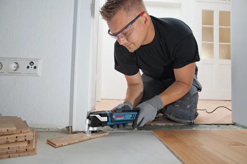 安全装備を身に着けた人が、出入り口の近くでマルチカッターを使用して床板をトリミングしています。.