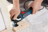 人がマルチカッター電動工具を使って床で木材を切断しています。.