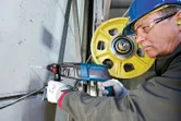 安全装備を身に着けた人が、黄色い滑車の近くで回転ハンマーを使ってコンクリートに穴を開けています。.