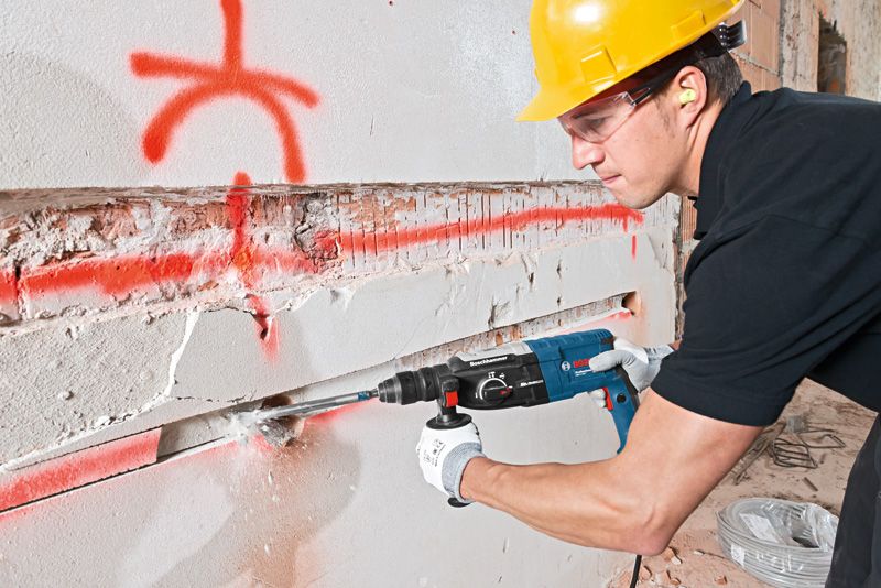 安全装備を身に着けた人が回転ハンマーを使用してコンクリートの壁に溝を彫っています。.