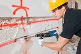 安全装備を身に着けた人が回転ハンマーを使用してコンクリートの壁に溝を彫っています。