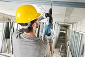 安全装備を身に着けた人が回転ハンマーで天井に穴を開けています。