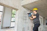 安全装備を身に着けた人が回転ハンマーを使用してコンクリートの壁に穴を開けています。