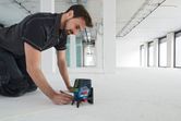 広い空き部屋で、レーザーレベル測定器を使って床に線を引いている人。
