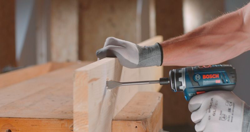 安全装備を身に着けた人が電動工具で木の梁に穴を開けています。