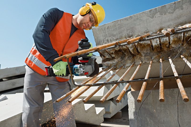 建設現場で、安全装備を身に着けた人が電動工具を使って突き出た鉄筋を切断している。.