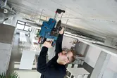 安全装備を身に着けた人が、近代的な作業スペースでコンクリートの天井にドリルで穴を開けています。.