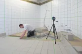 三脚に取り付けたレーザーレベル測定ツールを使用して、浴室にタイルを設置している人。