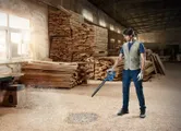安全装備を身に着けた人が、木工所で送風機を使っておがくずを吹き飛ばしている。
