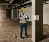 安全装備を身に着けた人が、建設現場でコンクリート柱にドリルで穴を開けている。