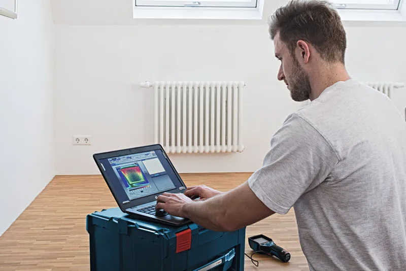 ラジエーターのある部屋でノートパソコンを使ってサーマルカメラのデータを分析している人。.