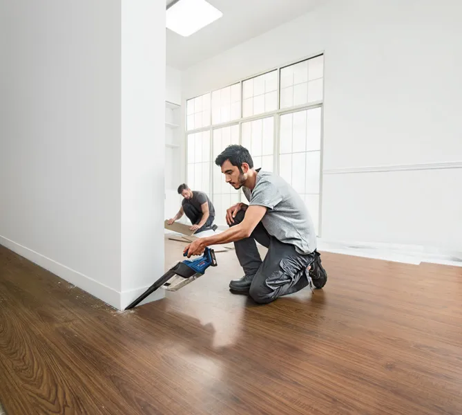 ある人がコードレスのこぎりで壁近くの床材を切断し、別の人が床パネルを設置しています。.