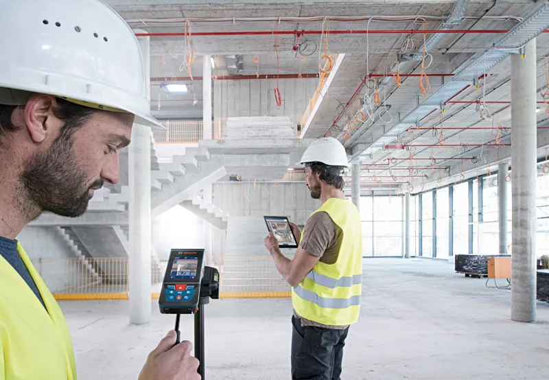 安全装備を身に着けた2人が、建設中の建物内でレーザー測定器とタブレット端末を使用している。