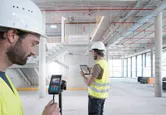 大規模な建設現場内で、安全装備を身に着けた2人がレーザー測定器とタブレットを使用しています。
