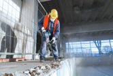 安全装備を身に着けた人が、解体用ハンマーを使用して屋内でコンクリートを破壊しています。