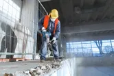 安全装備を身に着けた人が、解体用ハンマーを使用して屋内でコンクリートを破壊しています。.