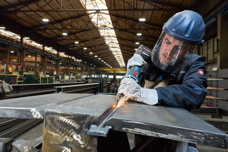 安全装備を身に着けた人が工場で電動工具を使って金属を研磨しています。.
