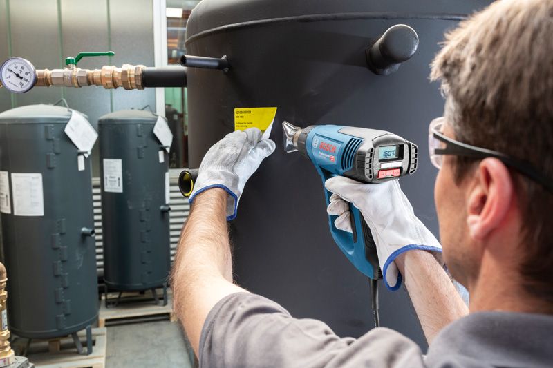 安全装備を身に着けた人がヒートガンを使用して大きなタンクにラベルを貼っています。