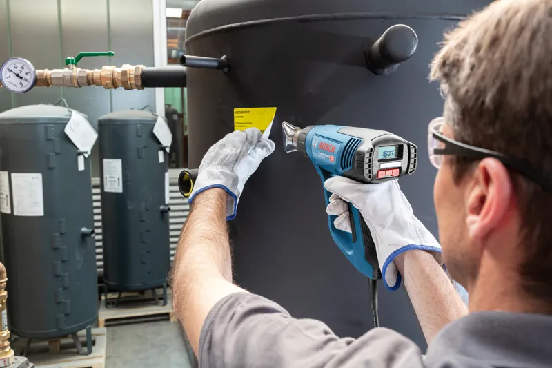 安全装備を身に着けた人がヒートガンを使用して大きなタンクにラベルを貼っています。