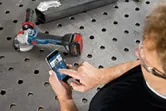 安全装置を装着した人がスマートフォンを使用して、金属作業台の上のコードレスグラインダーを制御します。
