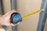 人が巻尺を使って乾式壁と金属フレームの間の距離を測っています。