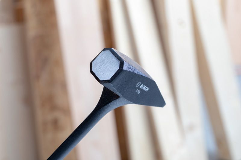 平らな打撃面を持つ黒いスレッジハンマーの頭が、ぼやけた木の背景に映っています。.