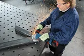 安全装置を装着した作業員が、穴あき作業台の上で金属の梁を研磨しています。