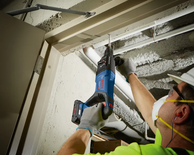 安全装置を装着した人が、コードレス回転ハンマーを使用してコンクリートの天井に穴を開けています。.