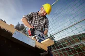 安全装置を装着した人が、建設現場でコードレス丸鋸を使用して木材を切断しています。