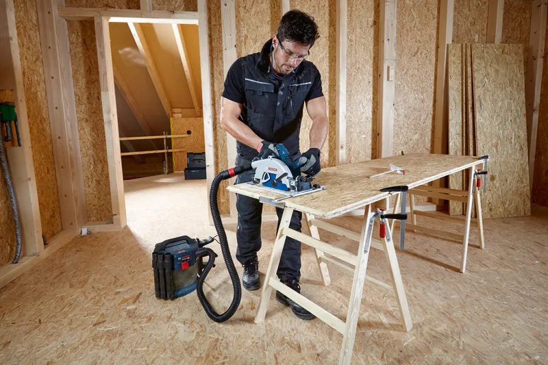 安全装備を身に着けた人が、集塵機に接続された丸鋸で合板を切断しています。.