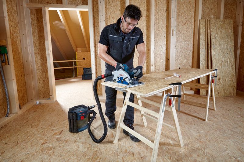 安全装備を身に着けた人が、集塵機に接続された丸鋸で合板を切断しています。.