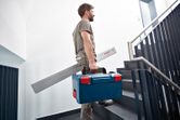 工具ケースと角度測定器を持ちながら階段を上る人。