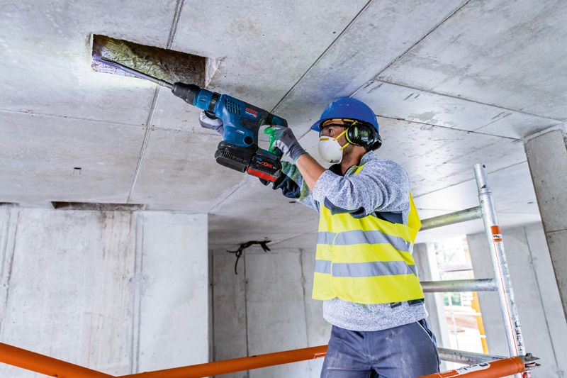 安全装備を身に着けた人がコードレス回転ハンマーでコンクリートの天井に穴を開けています。.