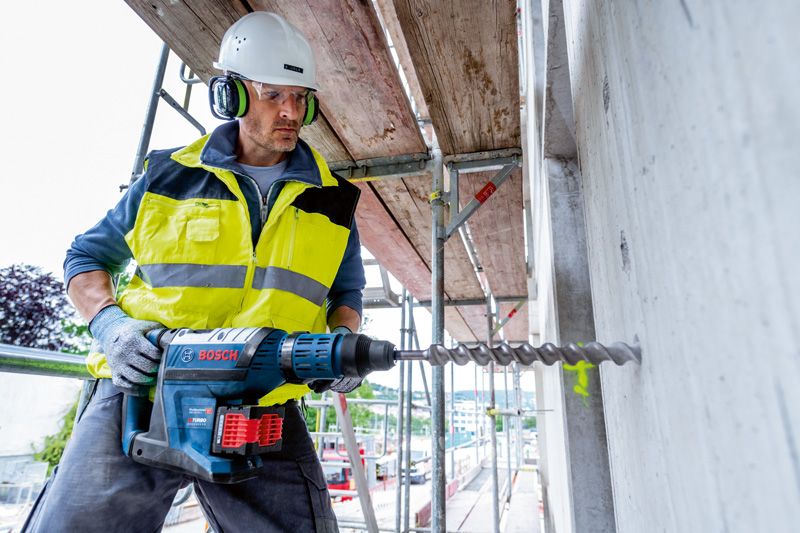 安全装置を装着した人がコードレス回転ハンマーでコンクリートに穴を開けています。.