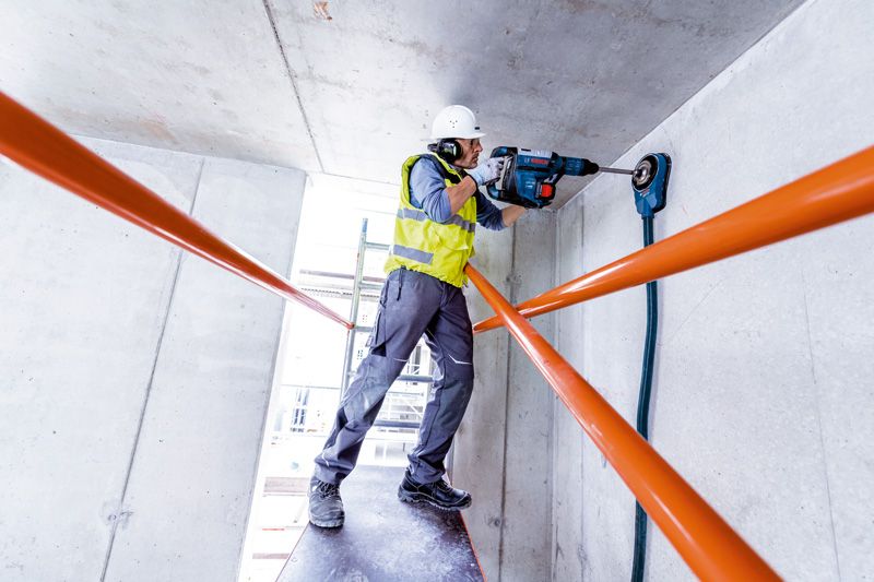 安全装備を身に着けた人が回転ハンマーでコンクリートの天井に穴を開けています。.