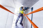 安全装備を身に着けた人が回転ハンマーでコンクリートの天井に穴を開けています。