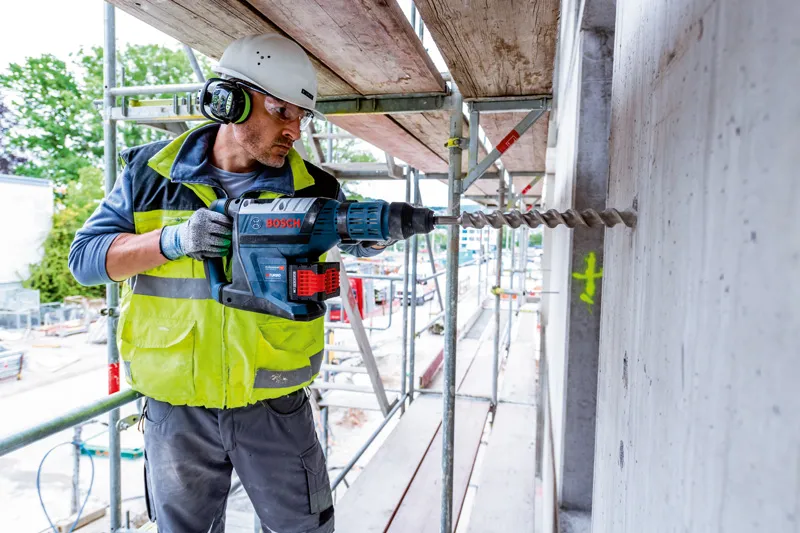 安全装備を身に着けた人が建設現場でコンクリートの壁に穴を開けています。.