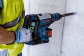 安全装備を身に着けた人が、大きな回転ハンマーでコンクリートに穴を開けています。