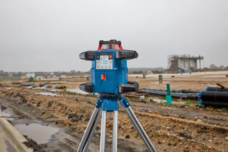 濡れた建設現場に三脚に取り付けられたレーザーレベル測定ツールが立っています。.