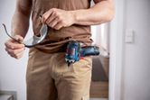 安全装置を装着した人がゴーグルを持ち、コードレスドライバーをベルトから下げています。
