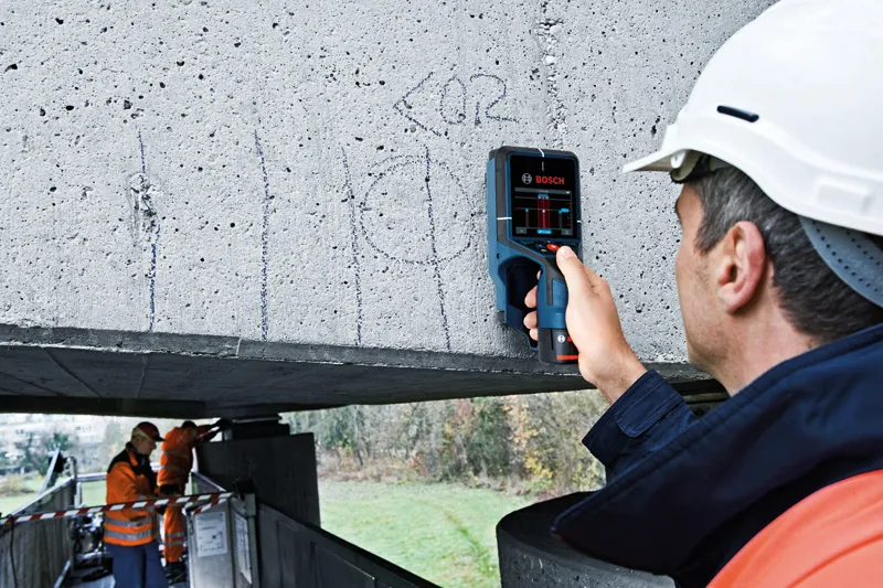 安全装備を身に着けた人が、検出器を使ってコンクリート梁をスキャンしている。