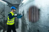 安全装備を身に着けた人が探知機でコンクリートの壁をスキャンし、隠されたパイプを発見する。