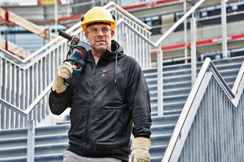 安全装備と加熱パーカーを着用した人が電動工具を持って階段に立っています。.