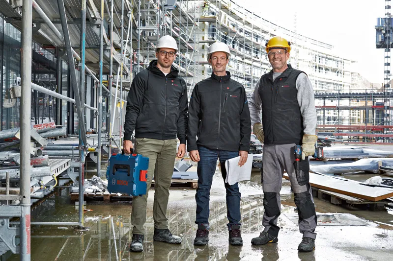 安全装備を身に着けた 3 人の作業員が建設現場に立っており、1 人は電動工具を持っています。.